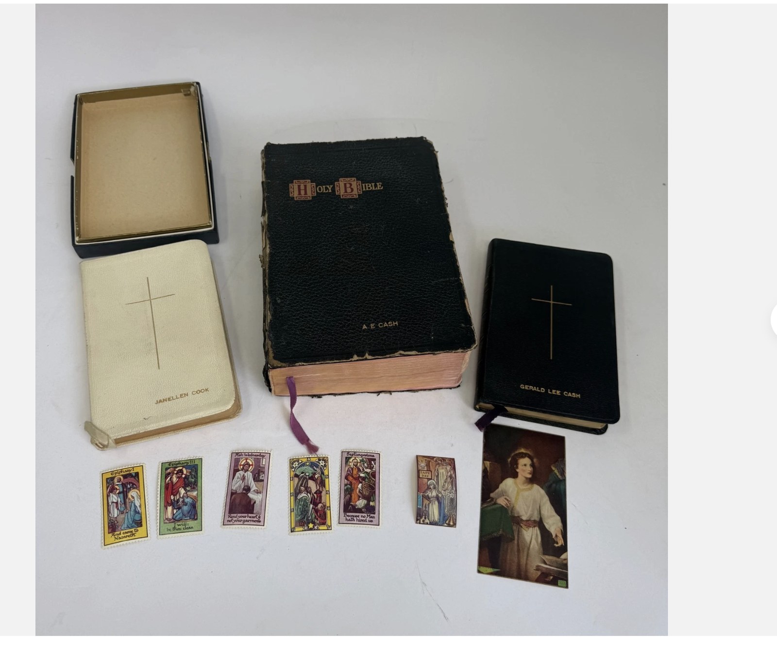 Three personalized family Bibles — a large leather Holy Bible stamped 'A. E. Cash', a white prayer book engraved 'Janellen Cook', and a black Catholic prayer book engraved 'Gerald Lee Cash' — shown alongside vintage religious prayer cards. The kind of family archive we handle for estate clients.
