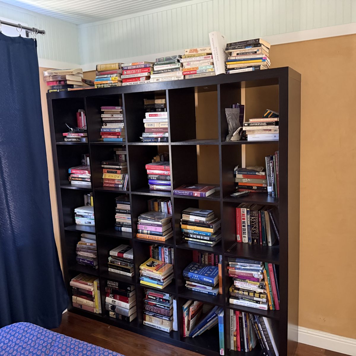 Dark wood Kallax-style bookshelf in a home, filled top-to-bottom with books, overflow stacks along the top shelf — a typical estate library before pickup.