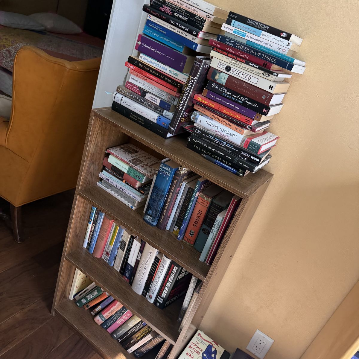 Mid-size home bookshelf with mixed hardcovers and paperbacks, a reading armchair visible in the background — the everyday lived-in library.