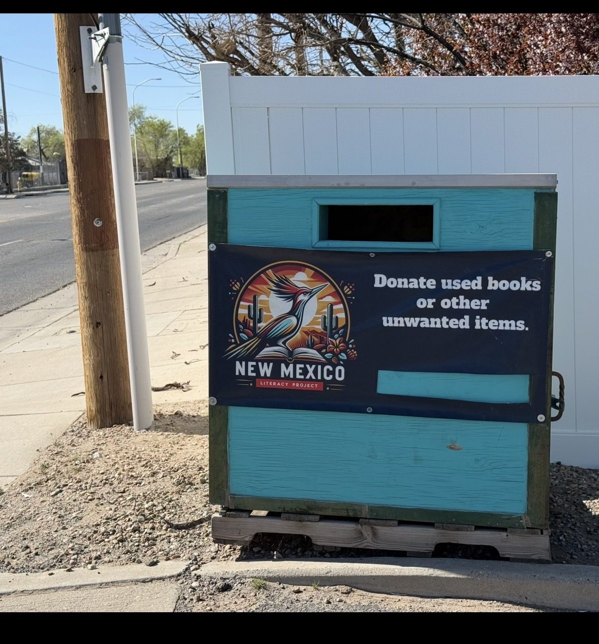 Teal wooden New Mexico Literacy Project donation drop box sitting on a pallet beside a sidewalk, with the roadrunner banner.